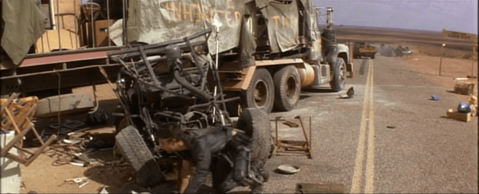 Max hunts out food and fuel from a wrecked truck, while the 'interceptor' is visible in the background.
